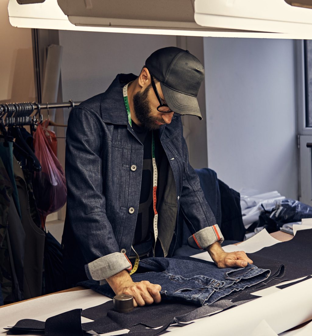 Handsome bearded tailor working with jeans material at a sewing workshop.